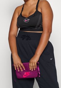 Svart sportbh med vit sömnad och rosa Nike-logotyp, i kombination med svarta sweatpants. Modellen håller en lila Nike-påse med rosa logotyp.