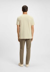 Man som står med ryggen mot kameran, iförd en beige kortärmad skjorta, bruna byxor och ljusa sneakers, mot en enfärgad background.