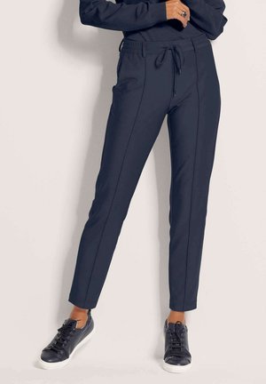 Vrouw draagt marineblauwe slim-fit broek met voornaad en koord in de taille, gecombineerd met zwarte sneakers en een bijpassende donkere top.