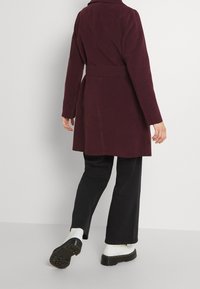 Abrigo burdeos con cintura ceñida, cuello con muescas y mangas largas, combinado con pantalones negros y botas de plataforma blancas, mostrado desde la parte posterior.