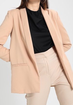 Femme portant un blazer beige et un pantalon avec un haut noir, debout les mains dans les poches devant un fond uni.