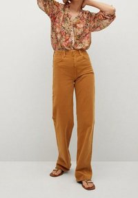 Vrouw in een bloeiende blouse met lange mouwen, hoge taille mosterdgele wijde broek en bruine teenslippers, staande tegen een effen achtergrond.