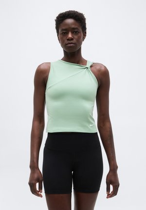 Žena s krátkymi kučeravými vlasmi, oblečená v svetlozelenom tielku bez rukávov s detailom na uzle a čiernych vysokých šortkách, stojaca na bielom pozadí.