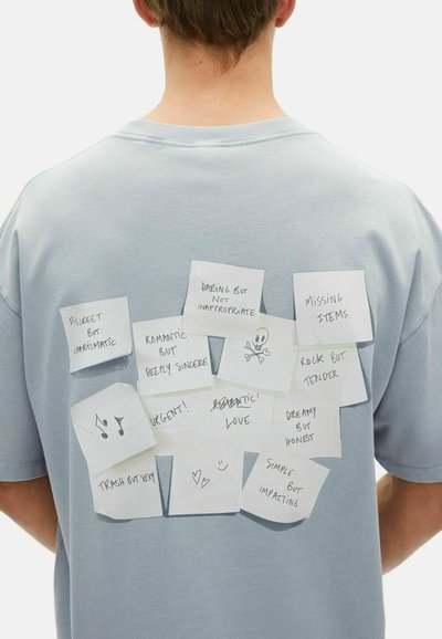 Camiseta de algodón azul claro con notas adhesivas escritas a mano con varias frases y bocetos pegados en la parte posterior. Diseño texturizado y casual.