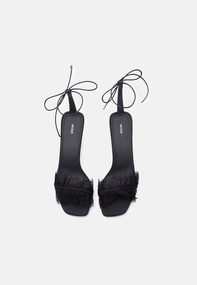 Sandales à talons hauts noires à bouts ouverts avec bordure en dentelle sur les brides et liens à la cheville, vues de dessus sur un fond blanc.