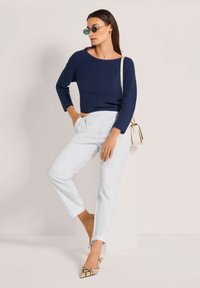 Blauer Strickpullover, hellblaue schmal geschnittene Hose und beigefarbene Schlangenleder-Pumps. Das Model ergänzt das Outfit mit runden Sonnenbrillen und einer kleinen Handtasche.