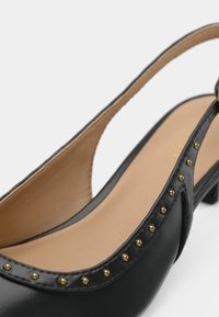 Chaussure noire à bride arrière avec une finition vernie brillante, bout arrondi, talon bas et des accents de clous dorés le long du bord. L'intérieur est beige.
