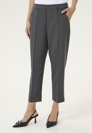 Kobieta ubrana w szare, dopasowane spodnie do kostek oraz czarne czółenka na obcasie typu slingback z ostrym noskiem, stoi z jedną ręką w kieszeni.