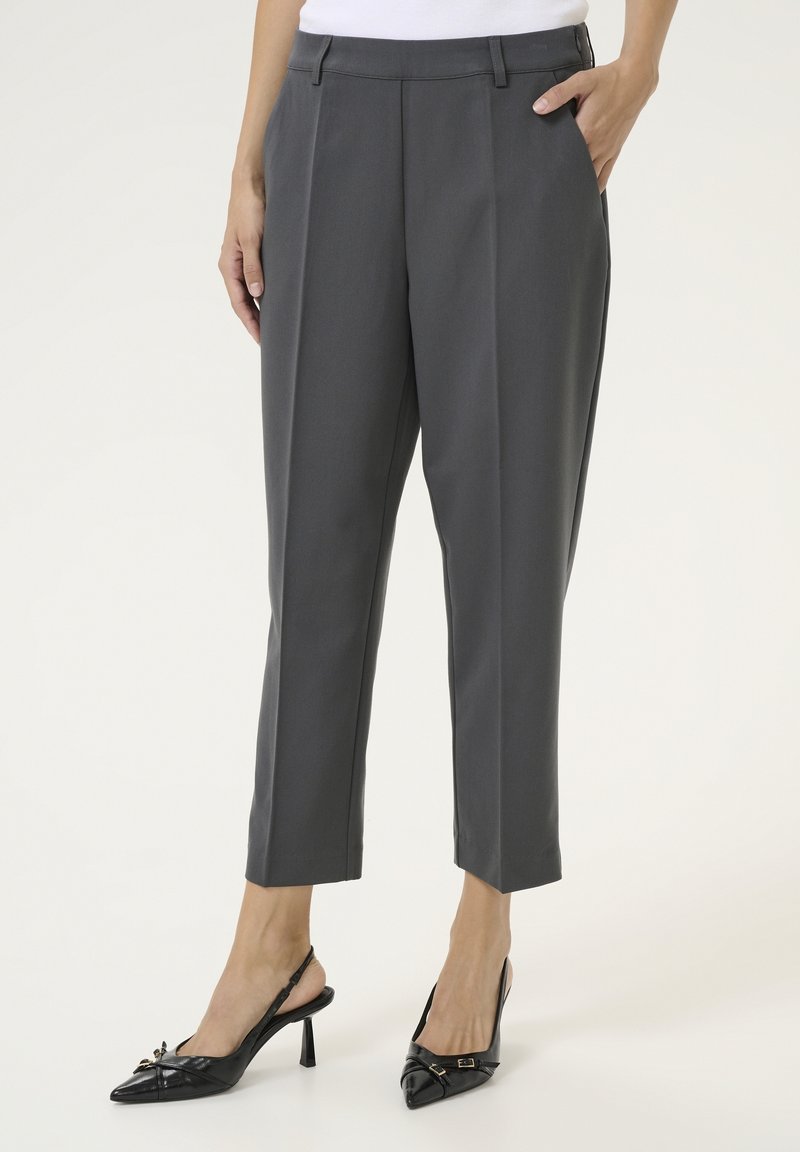 Femme portant un pantalon gris sur mesure longueur cheville et des escarpins slingback noirs à bout pointu, debout avec une main dans la poche.