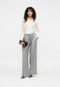 Valge seotav pluus pika varrukaga, millele on kombineeritud laiad hallid püksid ja mustad rihmikud sandaalid, hoides musta käekotti.