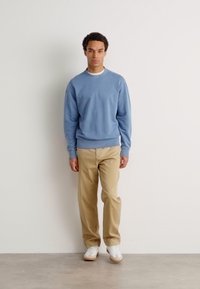 Ανοιχτό μπλε φούτερ με στρογγυλή λαιμόκοψη, μπεζ παντελόνι και λευκά αθλητικά παπούτσια. Το ύφασμα φαίνεται απαλό με άνετη εφαρμογή.