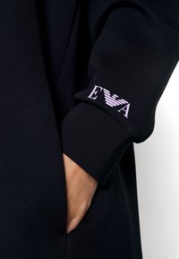 Schwarzer Sweatshirt mit weicher Textur, ausgestattet mit einer Tasche und einem lila EVA-Logo am Ärmel. Einfaches, minimalistisches Design.