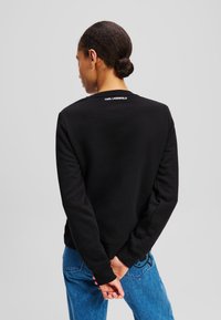 Sudadera negra fabricada con un tejido suave, presenta un cuello redondo y el logo de "KARL LAGERFELD" en la parte posterior, combinada con jeans azules.