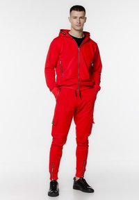 Czerwony dres składający się z bluzy z zamkiem błyskawicznym i spodni w stylu cargo. Wykonany z miękkiego materiału z czarnymi zamkami błyskawicznymi i detalami. Czernie sneakersy dopełniają wygląd.