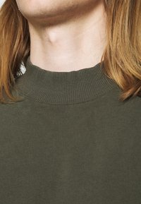 Suéter verde oscuro con un cuello alto acanalado y textura suave, con un diseño sencillo y sin patrones visibles ni adornos.