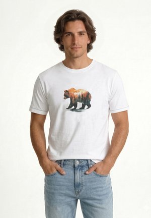 Muž so stredne dlhými vlasmi, ktorý má na sebe biele tričko s nákresom medveďa a lesa, stojí s rukami v vreckách a svetlomodrými džínsami.