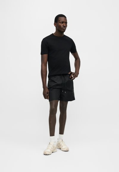 Άνδρας στέκεται με το χέρι στη μέση, φοράει μαύρο μπλουζάκι, μαύρες βερμούδες, λευκές κάλτσες και μπεζ αθλητικά παπούτσια μπροστά σε απλό ανοιχτό φόντο.