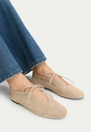 Piedi indossando ballerine in camoscio beige con lacci frontali, abbinate a jeans di jeans blu tagliati sopra la caviglia, su sfondo chiaro.