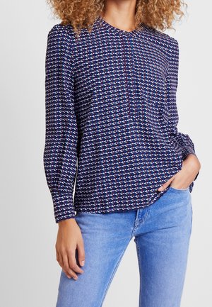 Kvinde iført en mønstret bluse med lange ærmer med geometriske figurer og lyseblå jeans, hånd i lommen, mod en ensfarvet baggrund.