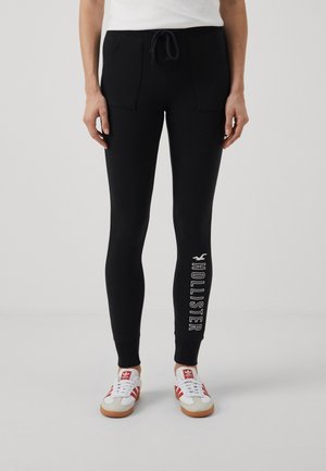 Person iført sorte tætsiddende joggingbukser med forlommer og teksten 'Hollister' på venstre nederste ben, kombineret med hvide og røde sneakers.