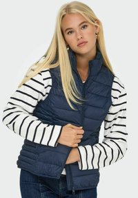Marine blauwe puffer vest met een hoge kraag, ritssluiting en een doorgestikt patroon, gedragen over een wit shirt met lange mouwen met marineblauwe strepen.