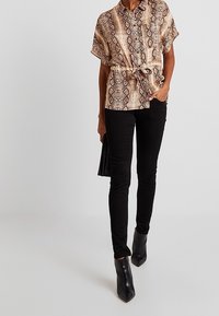 Blusa a maniche corte a motivi beige e marrone, cintrata in vita, abbinata a pantaloni skinny neri e stivali con punta alla caviglia.