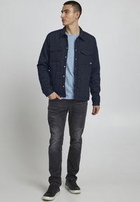 Marineblå vadderad jacka med två bröstfickor, knäppning fram, bärs över en ljusblå skjorta, kombinerad med mörka jeans och svarta sneakers.