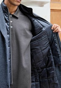 Palton texturat gri cu căptușeală neagră matlasată. Dispune de un fermoar și un guler cu nasturi. Sub el, se vede un tricou polo închis la culoare.