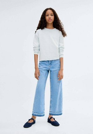 Giovane donna con capelli ricci vestita con un top a maniche lunghe grigio chiaro, jeans a gamba larga azzurri e scarpe Mary Jane blu scuro.