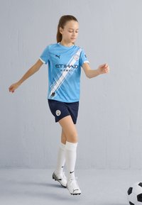 Camiseta de fútbol azul celeste con detalles blancos y escudo, combinada con pantalones cortos azul marino, calcetas blancas y botas de fútbol negras.