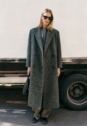 Femme aux cheveux blonds raides portant des lunettes de soleil noires, un long manteau gris texturé, un pantalon, des chaussures noires et tenant un sac à main noir à côté d’un véhicule.
