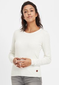 Langärmliger, cremefarbener Pullover mit rundem Ausschnitt, gerippten Bündchen und einem kleinen braunen Logoetikett am Saum. Weiches Strickmaterial.