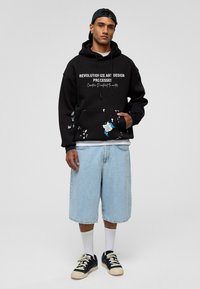 Zwarte hoodie met witte tekst, voorzien van een voorzak en verfpuntdetails. Combinatie met lichtblauwe denim shorts en zwarte sneakers.