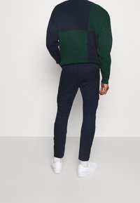 Grüner und marineblauer, farblich abgesetzter Strickpullover, schmal geschnittene marineblaue Hose mit zwei Gesäßtaschen, getragen mit weißen Sneakers.
