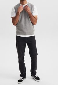 Grauer gerippter Strick-Sleeveless-Top über einem weißen T-Shirt, kombiniert mit dunklen Jeans und schwarzen Sneaker mit weißen Akzenten.