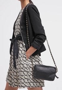 Blazer noir avec des accents en satin, robe blanche à motifs et un petit sac bandoulière en cuir noir avec une chaîne.
