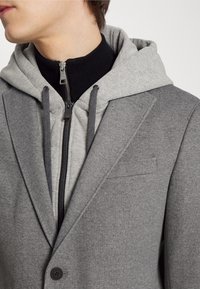 Mann trägt geschichtete graue Kleidung: einen hellgrauen Kapuzenpullover mit schwarzem Reißverschluss und Kordeln unter einem einmal geknöpften grauen Blazer.