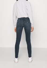 Person som bär mörkblåa tajta jeans, grå sweatshirt och vita höga Converse-sneakers, stående med ryggen mot betraktaren mot en enfärgad bakgrund.