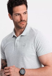 Camisa polo gris con un acabado texturizado, presenta un cuello con solapa, detalles de botones y un logotipo en el lado izquierdo del pecho. Reloj de pulsera con correa marrón visible.