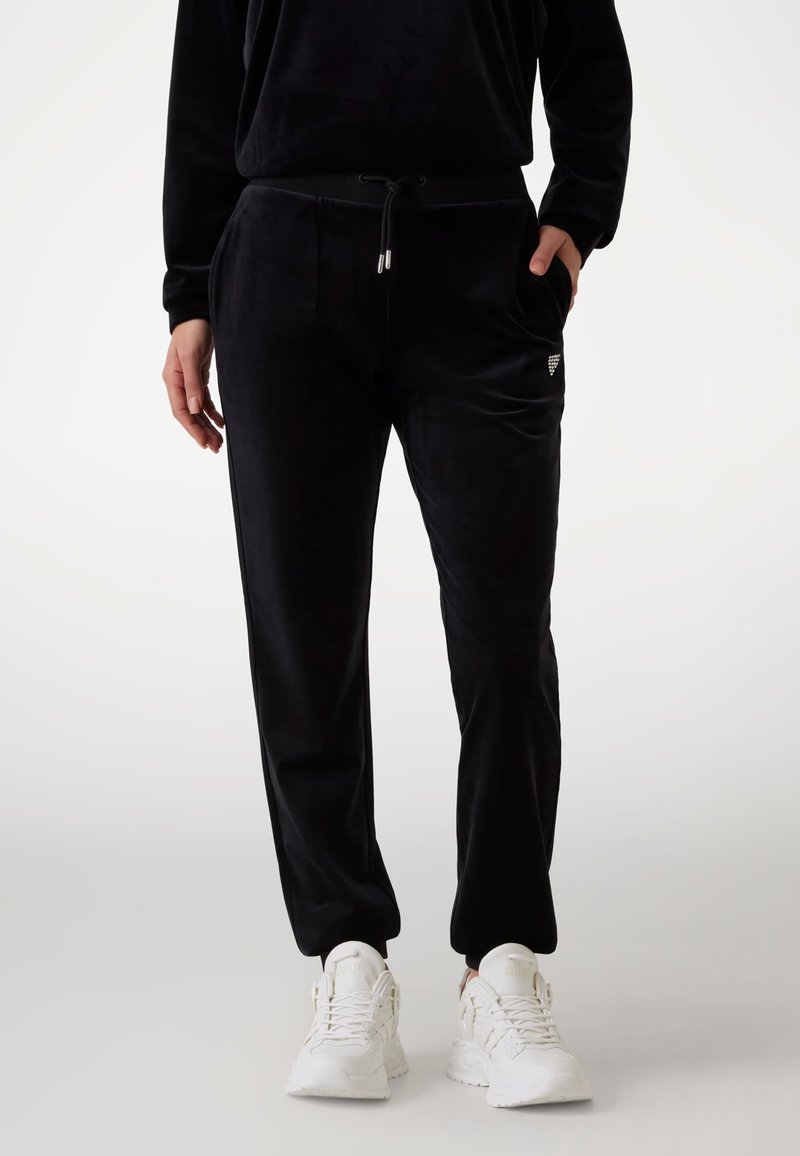 Svarte fløyels joggebukser med sidelommer, elastisk liv, og justerbar snor. Kombinert med hvite joggesko.