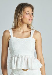 Top bianco a peplo con spalline larghe, ricami floreali testurizzati e orlo arricciato. Tessuto liscio con una leggera lucentezza; corpetto aderente.