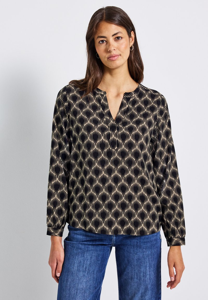 Donna con capelli lunghi e scuri, che indossa una blusa a maniche lunghe nera e beige con un motivo e jeans blu, in piedi davanti a uno sfondo semplice.
