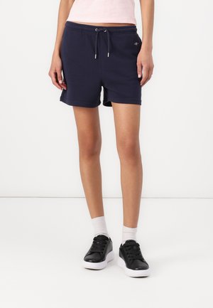 Vrouw draagt marineblauwe shorts met trekkoord, witte enkelsokken en zwarte sneakers, staand tegen een effen witte achtergrond.