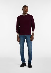 Maglione lavorato a maglia di un viola scuro con scollatura rotonda indossato sopra una camicia a quadretti bianca e verde, abbinato a jeans blu e scarpe nere.