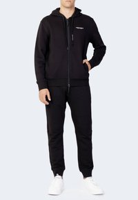 Hoodie zippé noir et joggers assortis en tissu doux, avec une poche frontale, des poignets côtelés et un détail de logo blanc sur la poitrine.