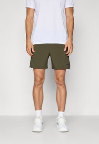 Calções verdes-oliva com um acabamento texturado, apresentando um corte reto e um pequeno logótipo preto na bainha inferior. Usados com uma t-shirt atlética branca.