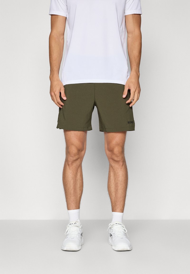 Calções verdes-oliva com um acabamento texturado, apresentando um corte reto e um pequeno logótipo preto na bainha inferior. Usados com uma t-shirt atlética branca.