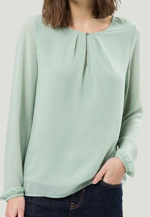 Kvinde iført lysegrøn bluse med lange ærmer, nøglehulshals og elastiske manchetter, kombineret med mørkeblå jeans.