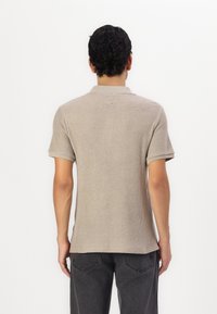 Polo à manches courtes, beige clair, présentant un design en maille texturée et un col classique. Porté avec un pantalon gris foncé.
