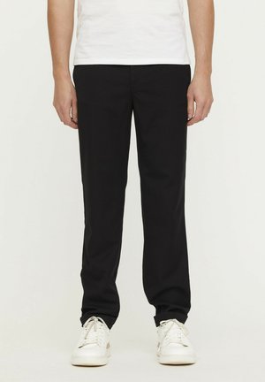 Pantalon noir en tissu tissé, avec une coupe droite, des poches avant et une texture lisse, associé à des baskets blanches.